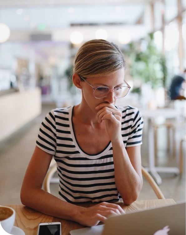 A person sitting at a café table, looking thoughtfully at a laptop screen with one hand resting near their mouth, suggesting concern or decision-making while working online.